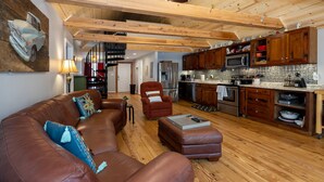 A spacious living room with a brown leather sofa, a red armchair, and a wooden ceiling. A kitchen with wooden cabinets and stainless steel appliances is visible in the background. - Awesome Lakefront House on West Point Lake in LaGrange, Ga (Lagrange)