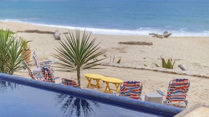 Un espace aménagé au bord de la piscine, avec des chaises longues et une table, offrant une vue sur une plage, une mer calme et une terre lointaine.