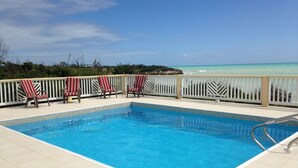 Una piscina con vista al océano, rodeada de sillones de descanso y una valla blanca.