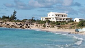 Una extensa propiedad frente a la playa, de color blanco, con varias construcciones, una playa de arena y aguas azules cristalinas.