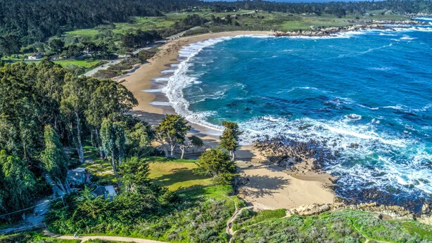 Vista aérea de uma área costeira com praia de areia, floresta densa e oceano azul cristalino.
