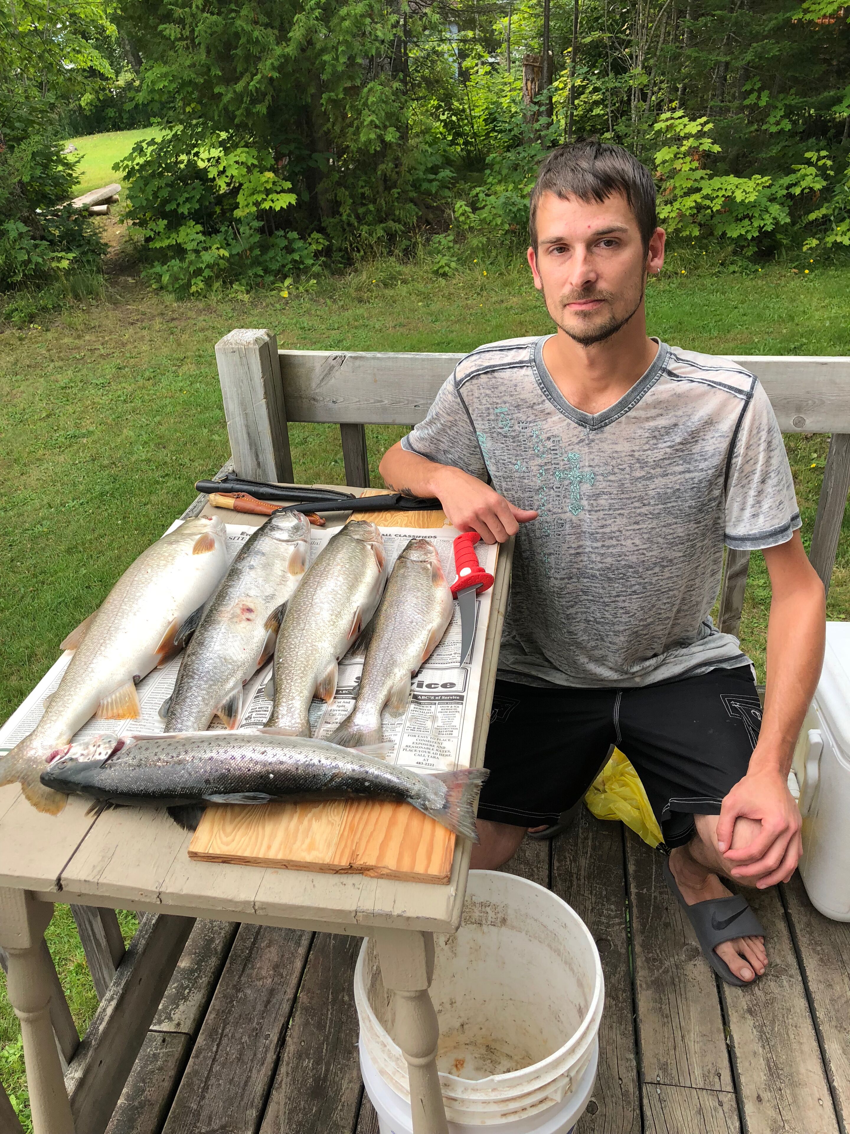 Une personne est assise à une table en bois, devant elle se trouvent plusieurs poissons posés sur du papier journal, un couteau et un seau.