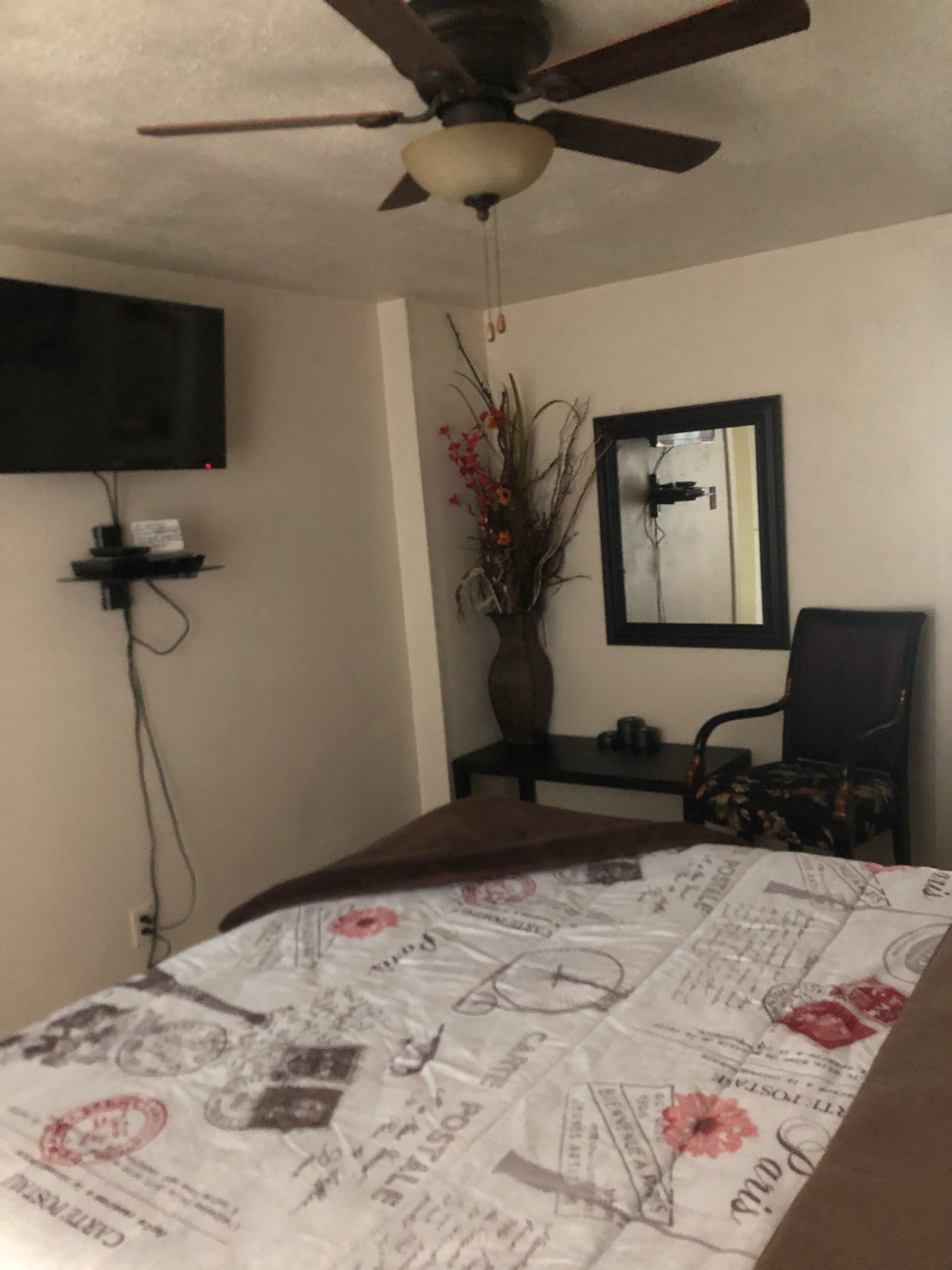 Ein Schlafzimmer mit Deckenventilator, einem Bett mit blumigem Überwurf, einem an der Wand montierten Fernseher, einer Vase mit Blumen und einem Sessel.