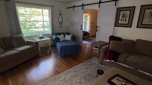 A living room with a sofa, ottoman, and a coffee table. There is a window with curtains, a lamp, and a door leading to another room. - Beautiful Private Harbert Beach cottage well appointed on a quiet private lane (Harbert)