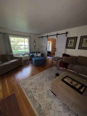 A living room with a sofa, ottoman, and a coffee table. There is a window with curtains, a lamp, and a door leading to another room.