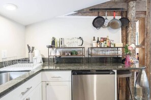 A kitchen with a dishwasher, a sink, a countertop with a dish rack, and a shelf with various kitchen items.