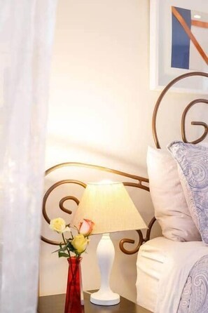 Una cama con cabecera, una mesita de noche con lámpara, y un florero con flores.