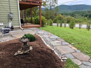 Un patio con un sendero de piedra, un pequeño jardín con un elemento decorativo de piedra, y una vista de un paisaje verde con árboles y una colina al fondo.