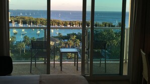 A balcony with a view of the sea, featuring a table and chairs, and a bed in the foreground.