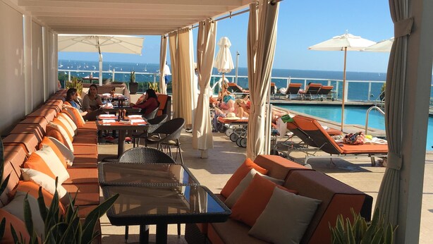 A covered outdoor seating area with orange and white cushioned sofas, a glass-top table, and potted plants.