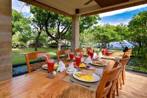 A dining table set with breakfast items and drinks, surrounded by chairs, in a covered outdoor area with a view of greenery and water. - Exotic Beachfront Villa in the Serene North (Pemuteran)