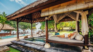 Un salon de bord de piscine doté d’un toit de chaume, d’une piscine bordée de pierres et d’une personne se relaxant sur un banc rembourré.
