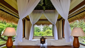 Une chambre avec un lit, deux lampes de chevet, un ventilateur de plafond et une vue sur une végétation luxuriante par la fenêtre.