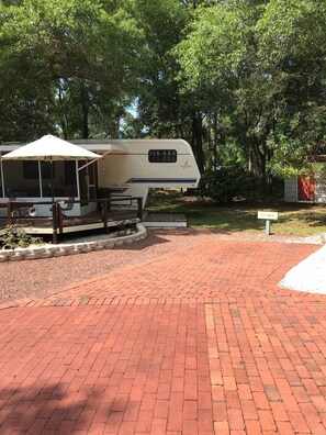 A covered RV with a patio area, surrounded by trees and a brick pathway. - Lets Go Camping In Comfort, Southern Style, come on down. (Summerdale)