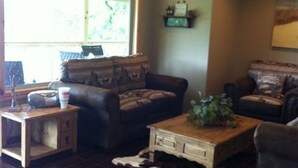 A living room with dark wood flooring, leather sofas, a wooden coffee table, and a large window with a view of greenery. - Most Beautiful View on Table Rock Lake! (Shell Knob)