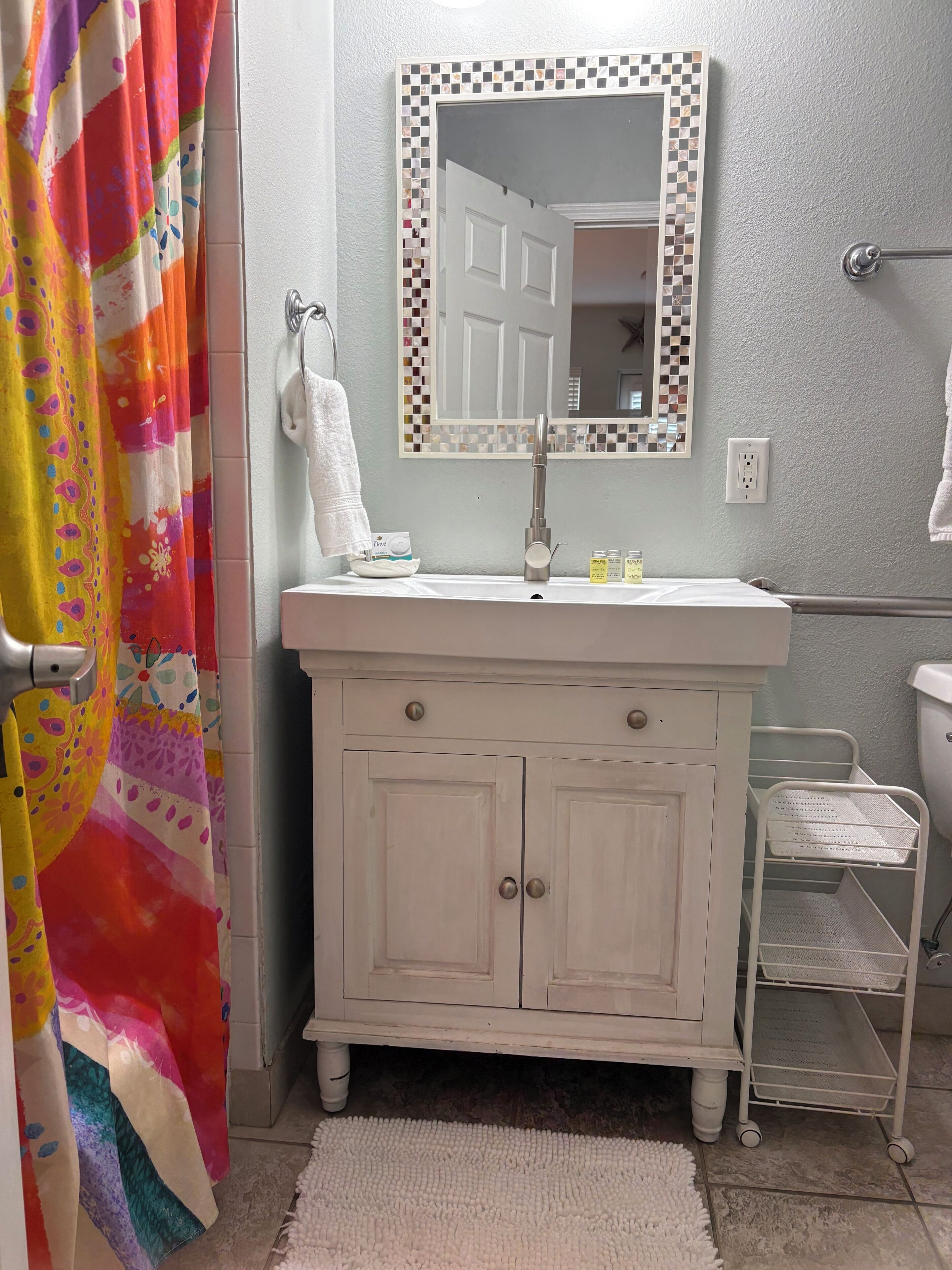 A bathroom with a white vanity, a mirror with a mosaic border, a shower curtain, and a white towel on the wall.
