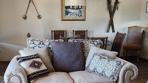 Un salon chaleureux avec un canapé beige, un pouf marron à motifs et un panneau d’accueil encadré.