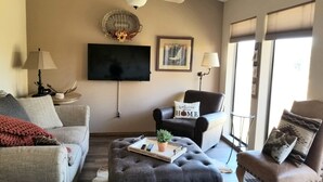 Un salon comprenant un canapé, un fauteuil, un pouf et un téléviseur à écran plat fixé au mur.