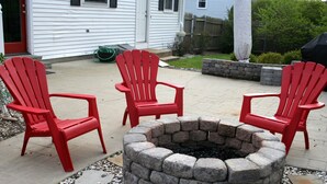 A fire pit surrounded by three red Adirondack chairs in a backyard with a stone wall and a white house in the background. - PRIVATE HOME NEAR BEACH LOCATED IN NARRAGANSETT, RI (Narragansett)