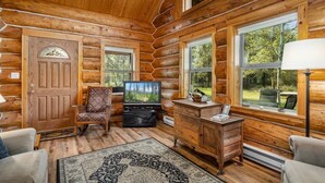 A log cabin interior with a wooden door, a television, a chair, a desk, and large windows. - Steps to the River! River Edge Cabin (West Glacier)