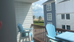 Un balcon avec deux chaises Adirondack bleues et une table, donnant sur une plage et un bâtiment aux murs recouverts de panneaux blancs et bleus.