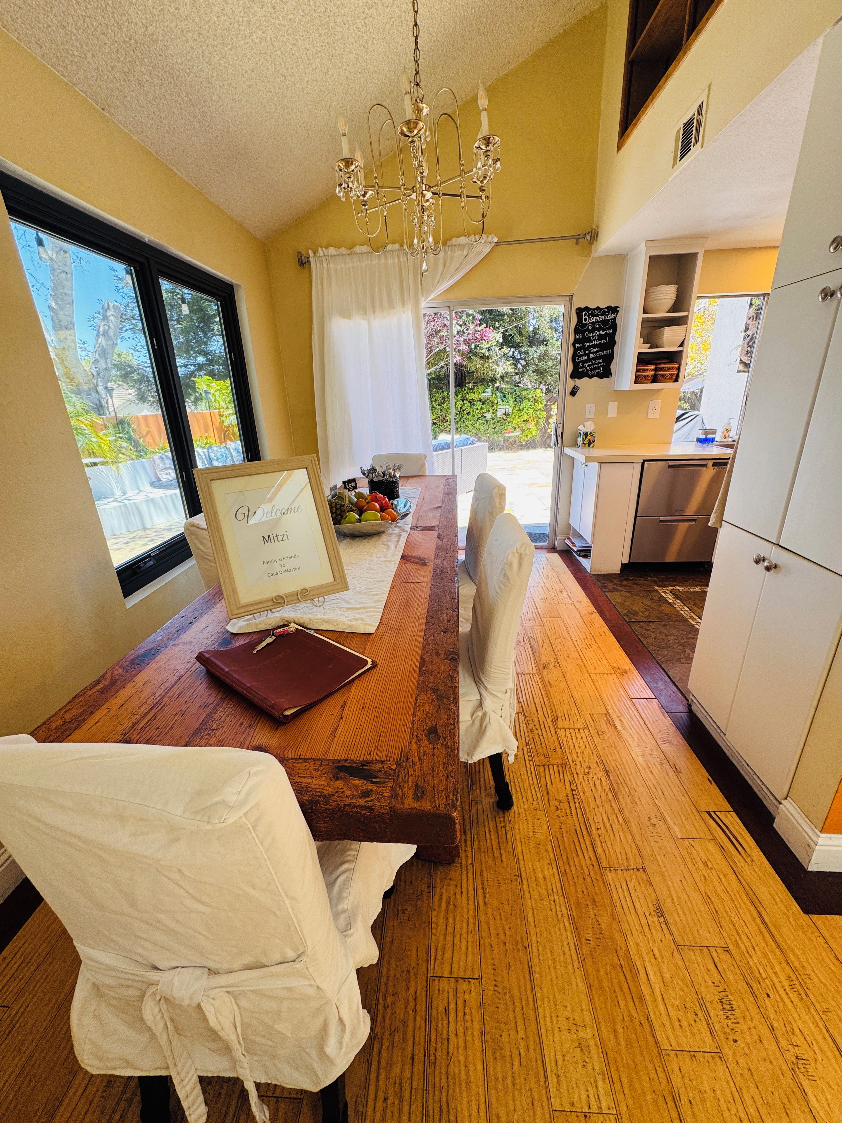 A dining area with a wooden table, white chairs, a chandelier, and a view of the garden through the window.