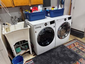 Zona de bugaderia amb dues rentadores, un bany i un cub blau de plàstic amb productes de neteja.
