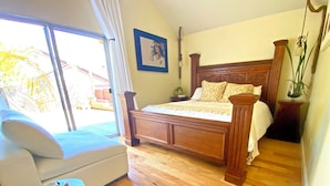Une chambre avec un lit en bois, un canapé blanc, une petite table d’appoint blanche et une grande fenêtre donnant sur des palmiers.