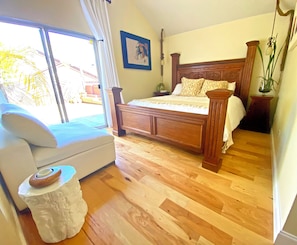 Une chambre avec un lit en bois, un canapé blanc, une petite table d’appoint blanche et une grande fenêtre donnant sur des palmiers.