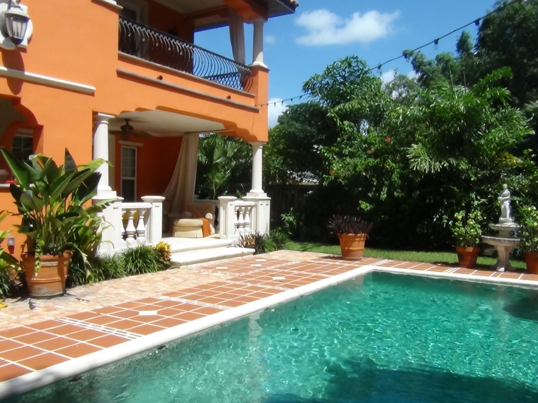 Une piscine dont le bord est recouvert de carreaux, entourée de plantes en pot et bordée d’un immeuble à deux étages doté d’un balcon.