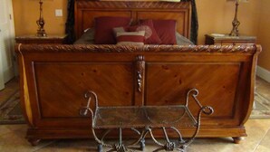 Een houten bed met een metalen hoofdbord en voeteneind, een nachtkastje met een lamp, en een raam met gordijnen.