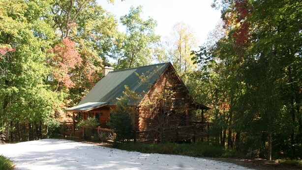 A rustic log cabin with a green roof, surrounded by trees and a gravel driveway.