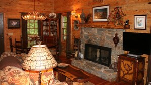 A cozy log cabin interior with a stone fireplace, a television, and a dining area with chairs and a table.