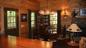 A wooden cabin interior with a dining table, chairs, a chandelier, and a fireplace.