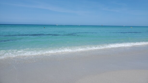 Una playa con agua turquesa cristalina, orilla arenosa y barcos a lo lejos.