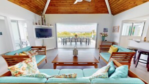 Uma sala de estar ampla com teto de madeira, uma TV grande, um espaço de jantar com assentos externos e vista para o exterior.