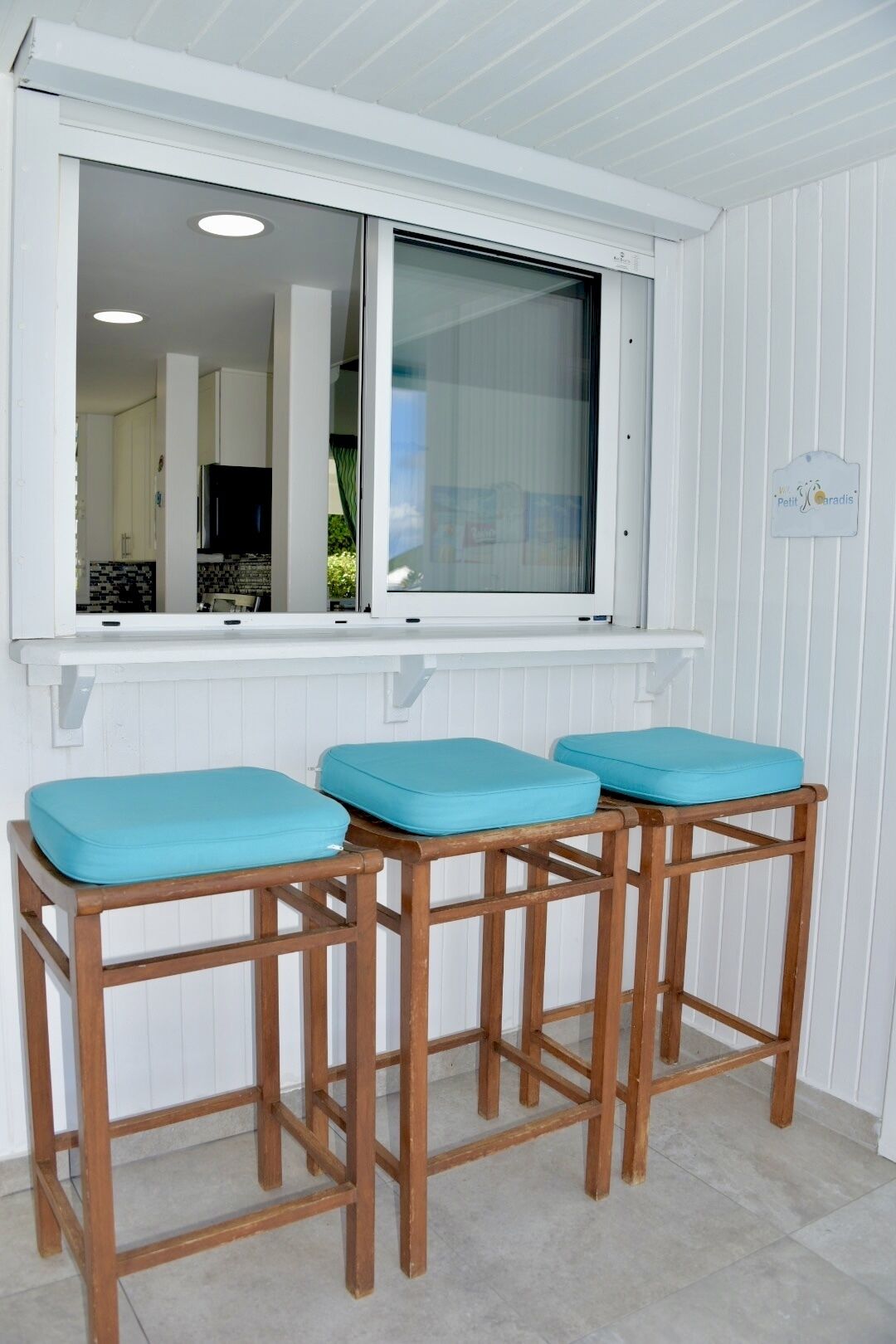 A kitchen with white cabinets, a window with a view of the interior, and a wooden stool with a blue cushion.