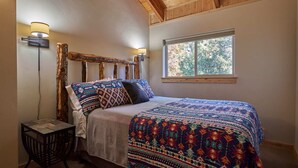 Ein Schlafzimmer mit einem Holz-Kopfteil, einer gemusterten Bettdecke, einem Nachttisch und einem Fenster mit Blick auf Bäume.