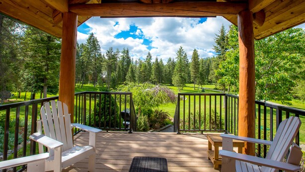 Una terraza de madera con dos sillas Adirondack blancas y un banco, con vistas a un paisaje verde exuberante con árboles y un campo de golf.