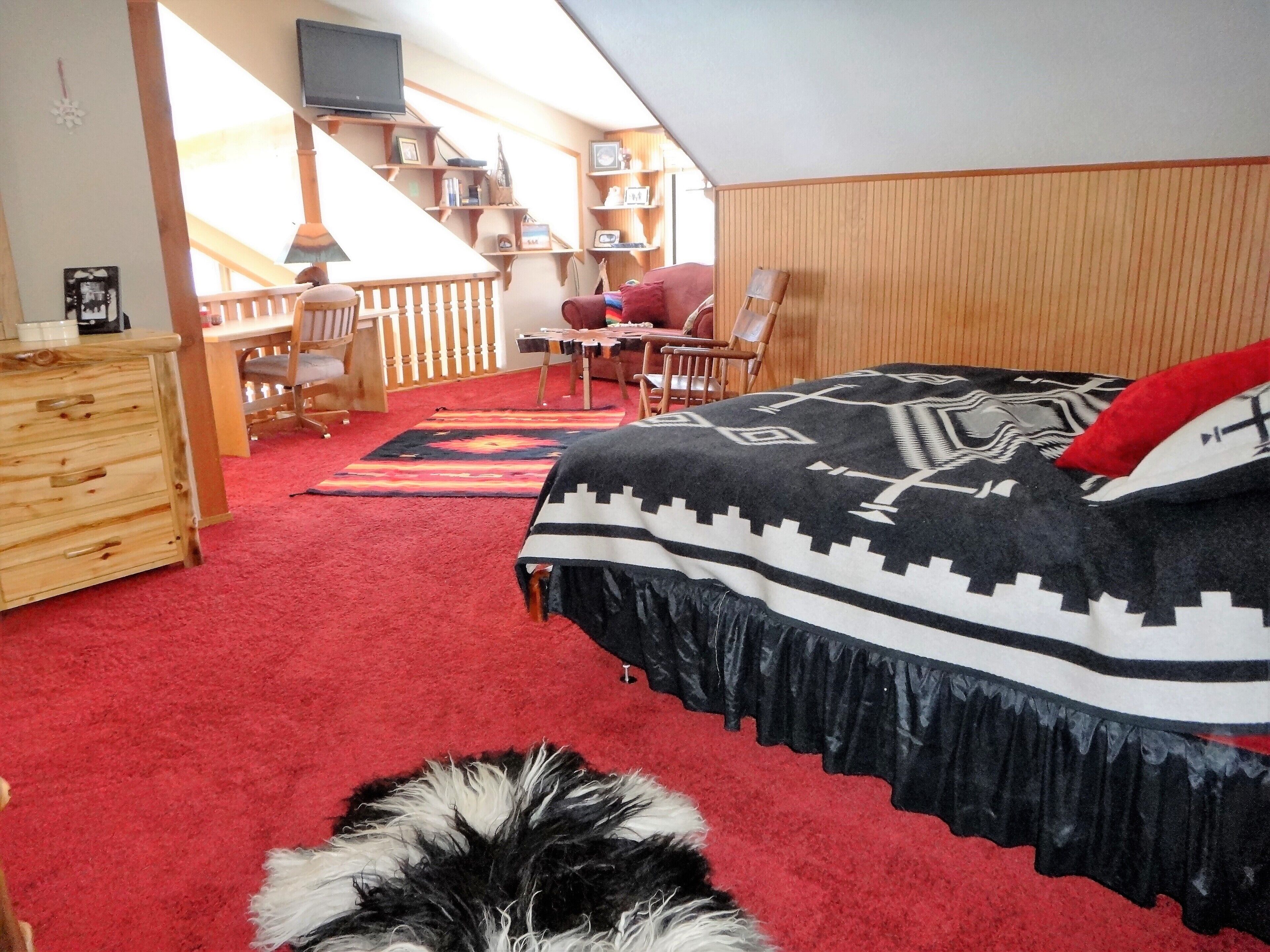 Une chambre avec une moquette rouge, un lit recouvert d’une couverture à motifs, une commode en bois, un bureau avec une chaise et un téléviseur fixé au mur.