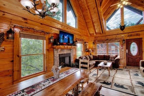 A cozy cabin interior with a fireplace, wooden furniture, large windows, and a ceiling fan.