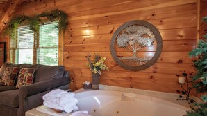 A wooden cabin interior with a large bathtub, a couch with patterned pillows, and a wall-mounted tree decoration. - A+ Views & Location! Minutes 2 Dollywood. Hot Tub Games Pet Friendly Resort Pool (Pigeon Forge)