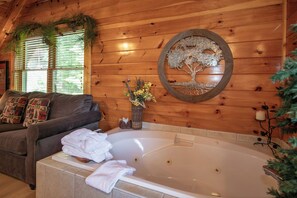 A wooden cabin interior with a large bathtub, a couch with patterned pillows, and a wall-mounted tree decoration.