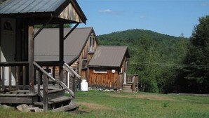 Une cabane rustique dotée d’une terrasse en bois et d’un toit en métal, entourée d’une zone herbeuse et bordée, au loin, par une colline boisée.