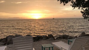 Une plage avec des chaises longues blanches et une petite table, un rivage rocheux et un voilier à l’horizon, au moment du coucher du soleil.