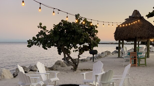 Un espace de détente aménagé sur la plage, avec des fauteuils Adirondack blancs, un foyer en pierre et des guirlandes lumineuses.