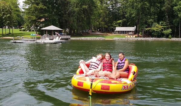A group of people on a yellow inflatable raft on a lake, surrounded by trees and a dock with a covered structure. - Lake Front Getaway, Family Friendly. (Mooresville)