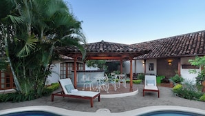 Zona de piscina con una pérgola, sillones de descanso y un patio empedrado.