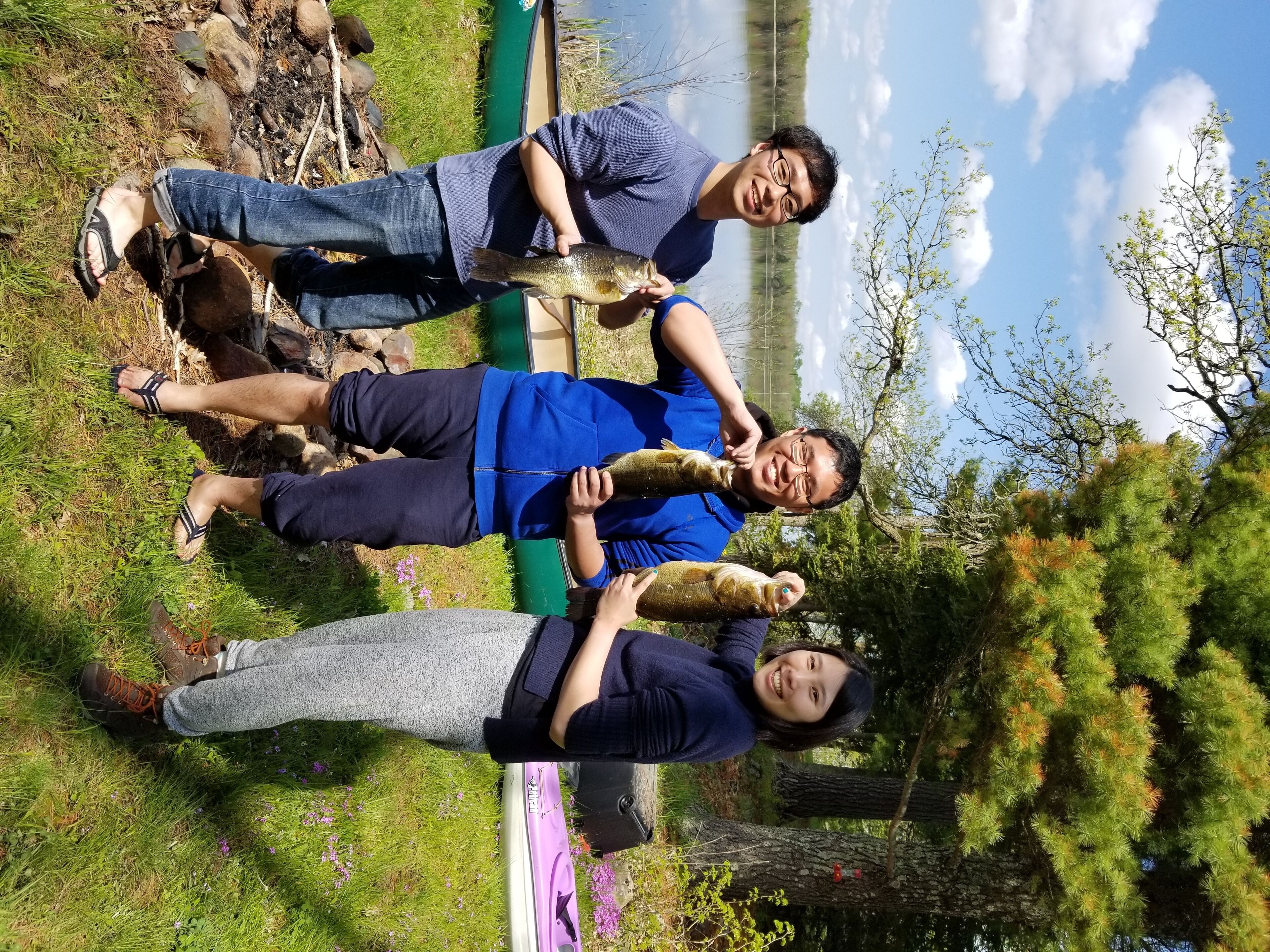 Trois personnes se trouvent à l’extérieur, près d’une étendue d’eau, et l’une d’entre elles tient un poisson.