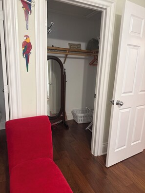 Una habitación con una silla roja, suelo de madera, una puerta blanca, un espejo, un cesto de ropa y un perchero.